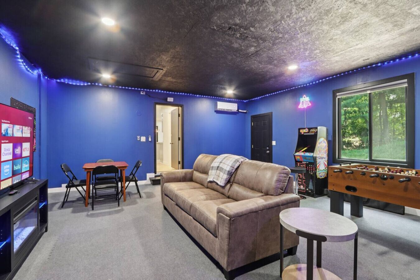 A room with a blue wall, a leather couch, a foosball table, a TV, and a small table with chairs.
