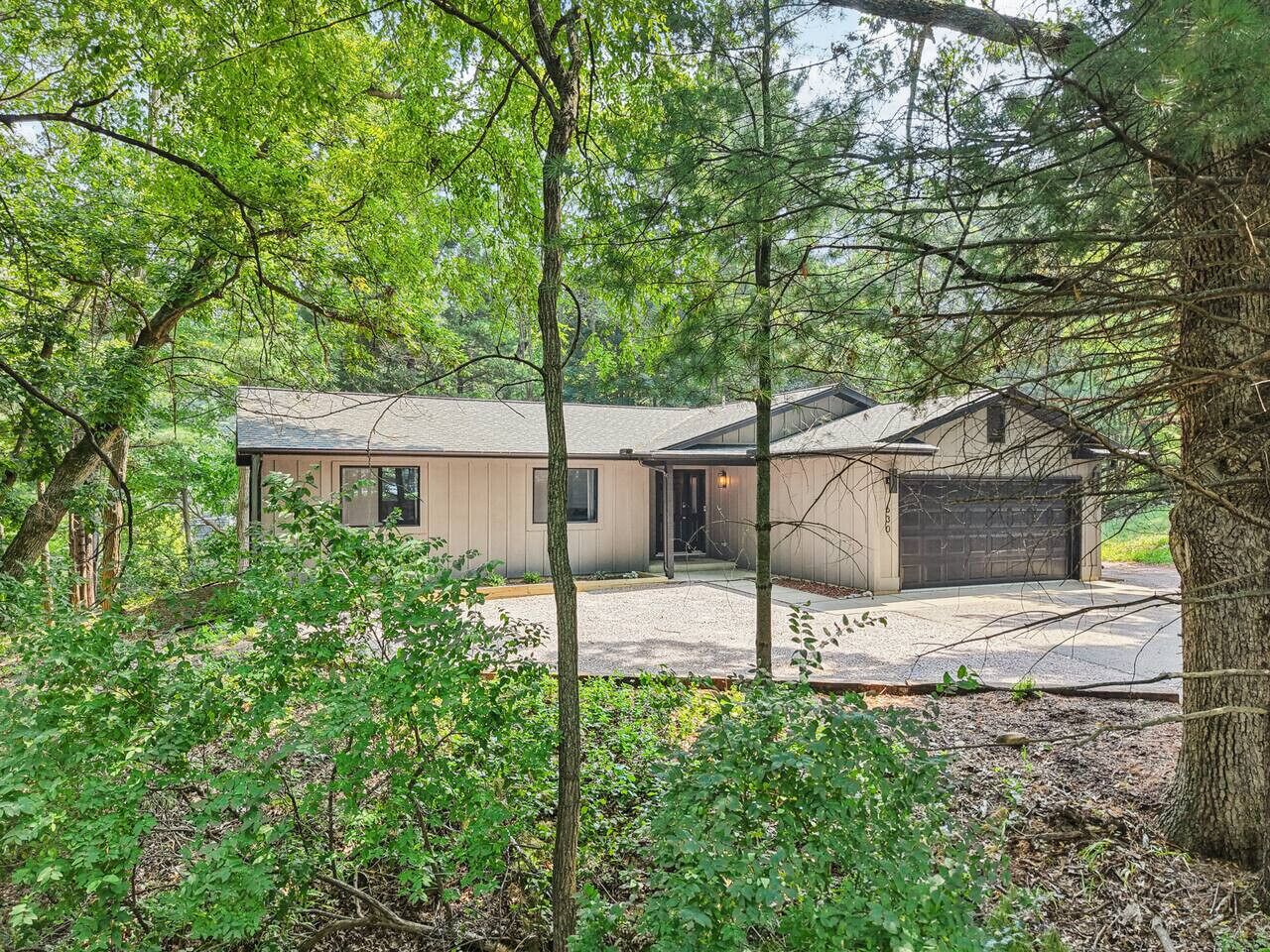 Une maison moderne à deux étages avec un garage, entourée d’arbres denses et dotée d’une allée en gravier.