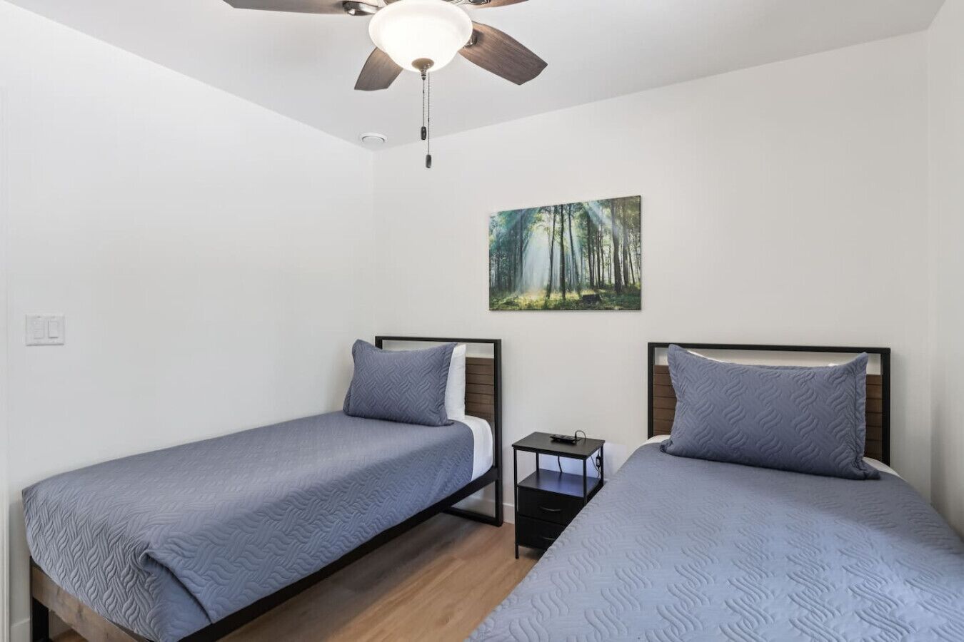 A bedroom with two single beds, a ceiling fan, a nightstand, and a painting of a forest.