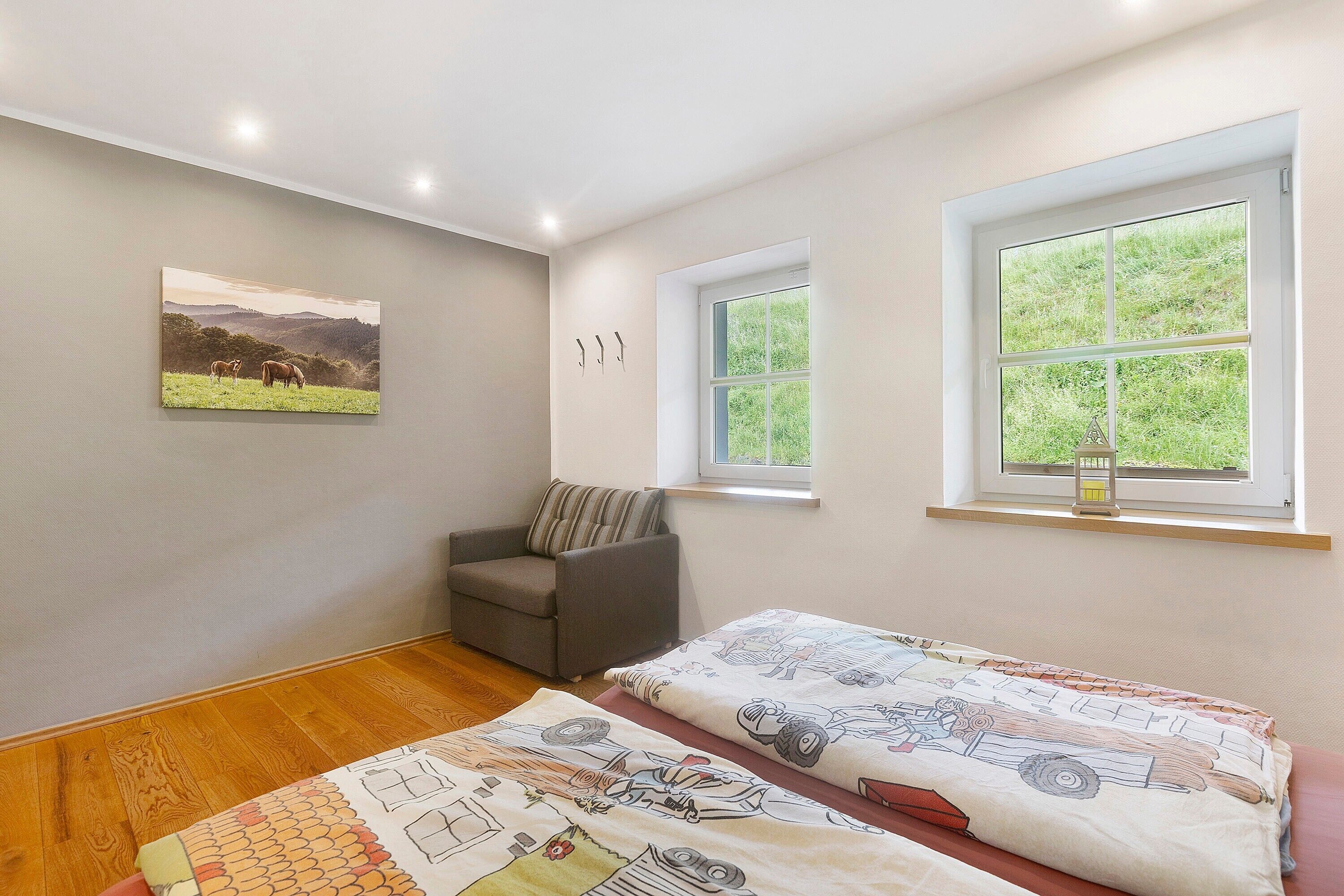 A room with a bed, a sofa, a window with a view of greenery, a painting on the wall, and a small table with a lantern.