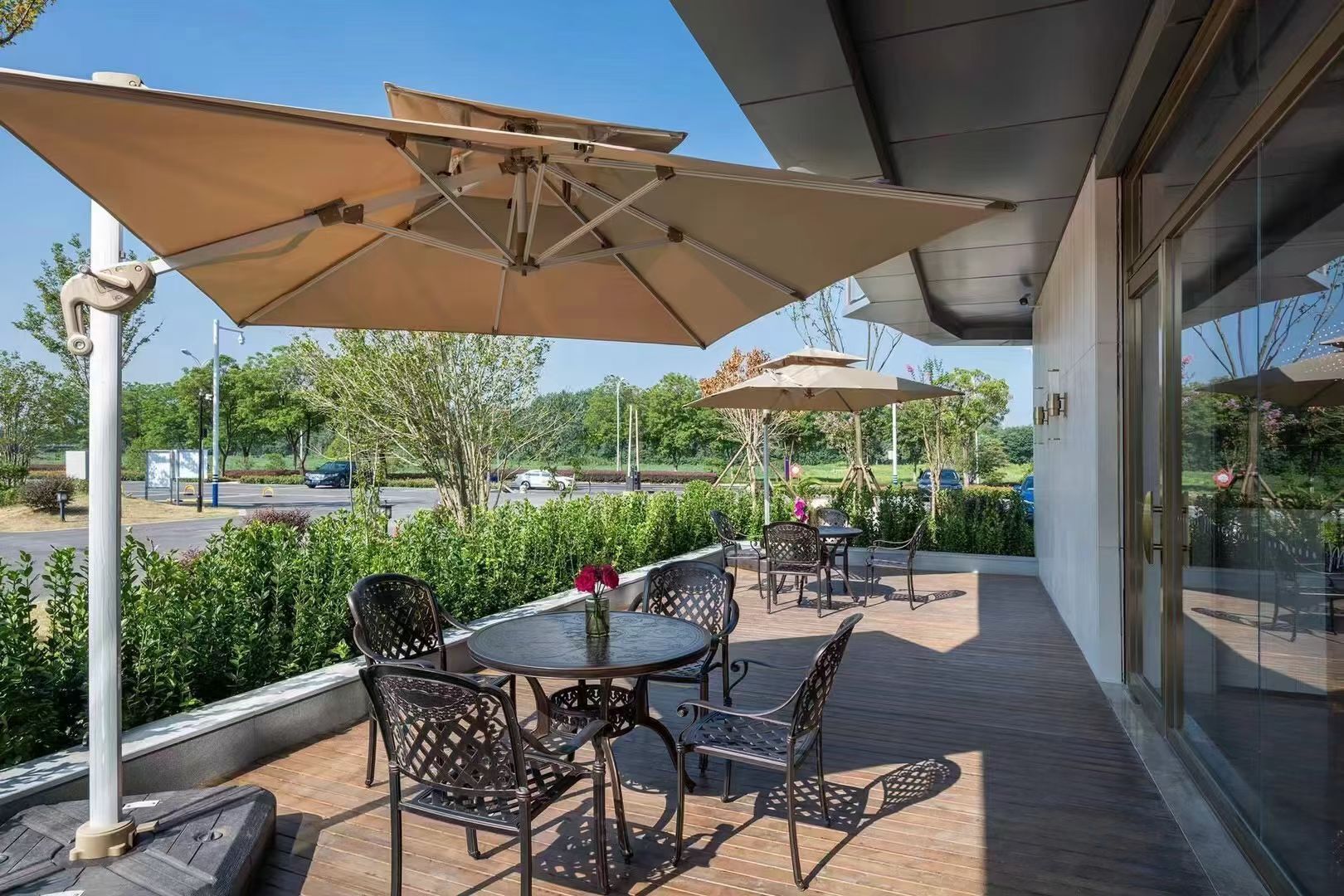 En udendørs terrasse med borde, stole, parasoller og udsigt til parkeringsplads og grøn bevoksning.