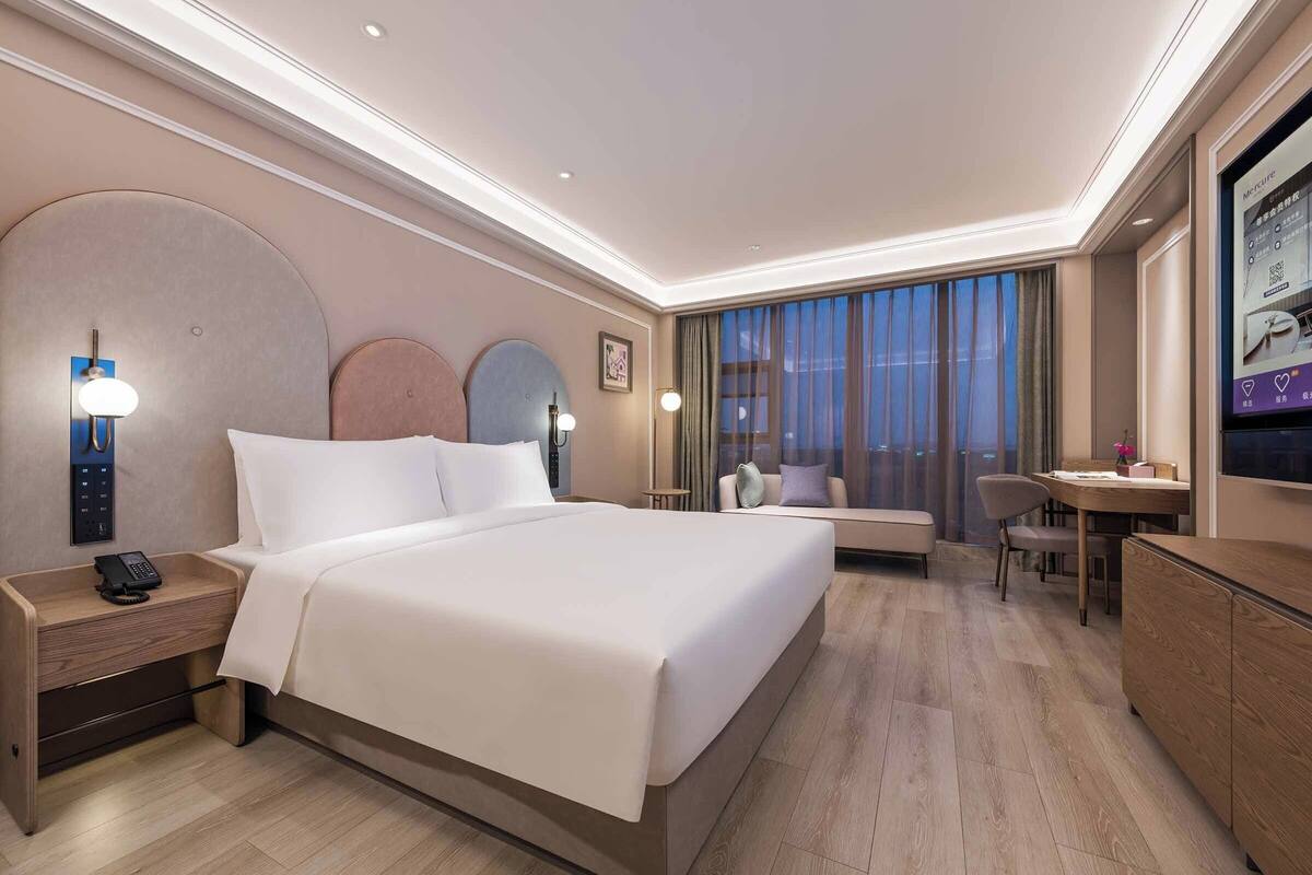 A hotel room with a large bed, bedside table with a phone, a desk with a chair, and a large window with curtains.