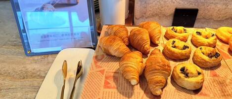 A breakfast buffet with croissants and other pastries, a digital menu board, and a plate with cutlery.