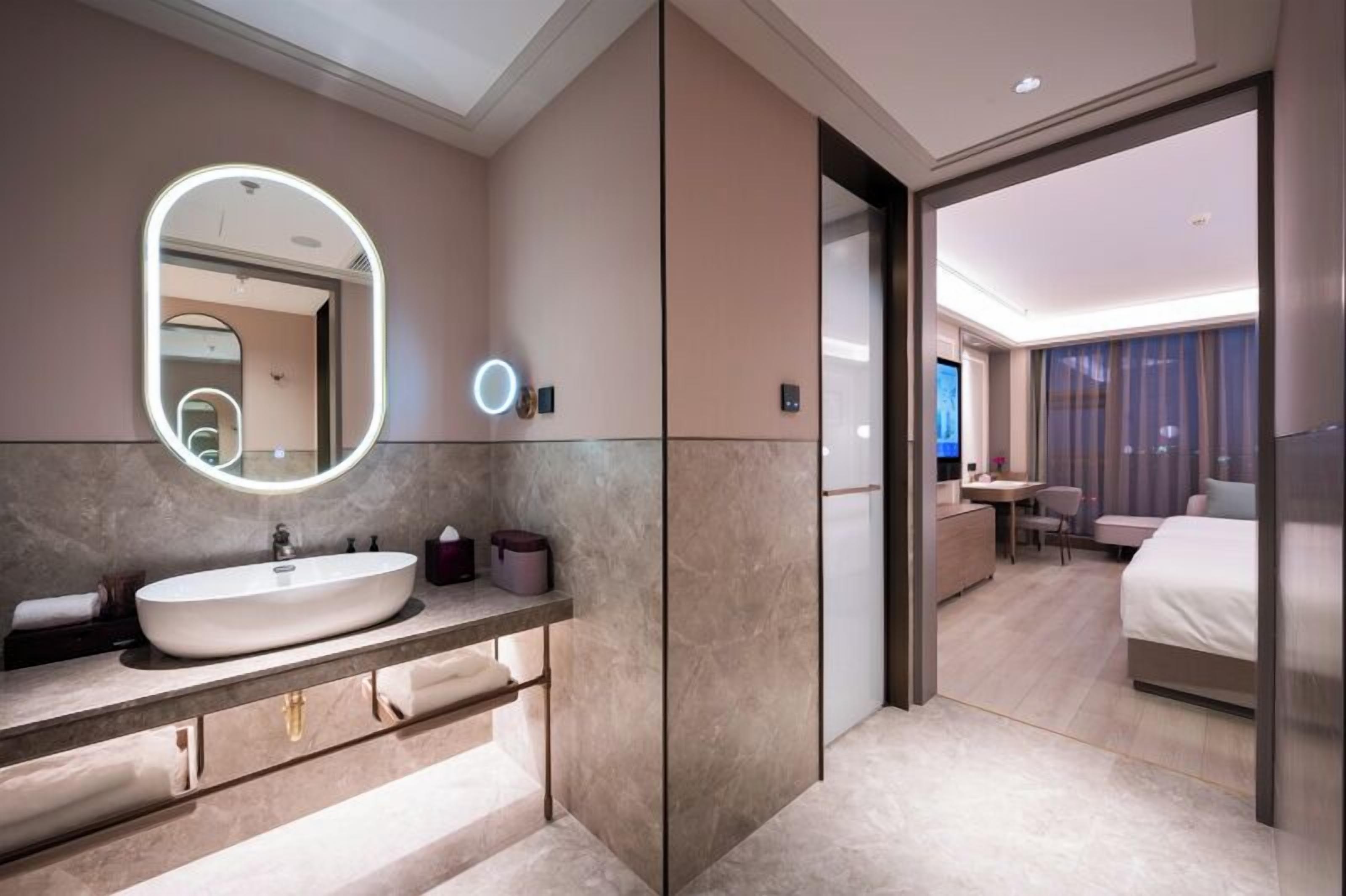 A modern hotel bathroom with a large oval sink, a mirror with a light, and a view into the bedroom.
