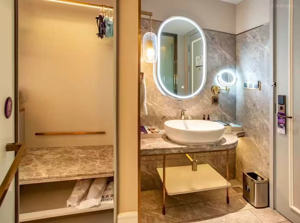 Un baño con un espejo ovalado grande, un lavamanos blanco, un perchero y un cubo de basura.