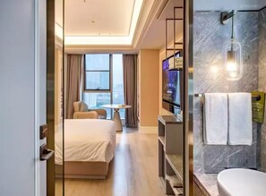 Hotelkamer met een bed, een bureau, een stoel, een klein tafeltje, een badkamer met wastafel en spiegel, en uitzicht op de stad door grote ramen.