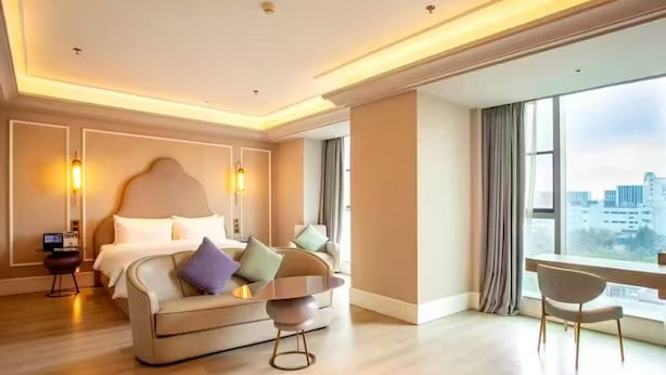 Habitación de hotel moderna con una cama grande, un sofá, una mesita y vistas de la ciudad a través de amplias ventanas.