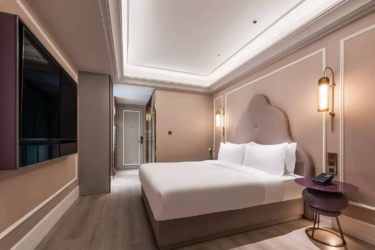 A hotel room with a large bed, bedside table, wall-mounted TV, and a hallway leading to another room.