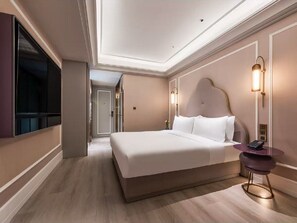 A hotel room with a large bed, bedside table, wall-mounted TV, and a hallway leading to another room.