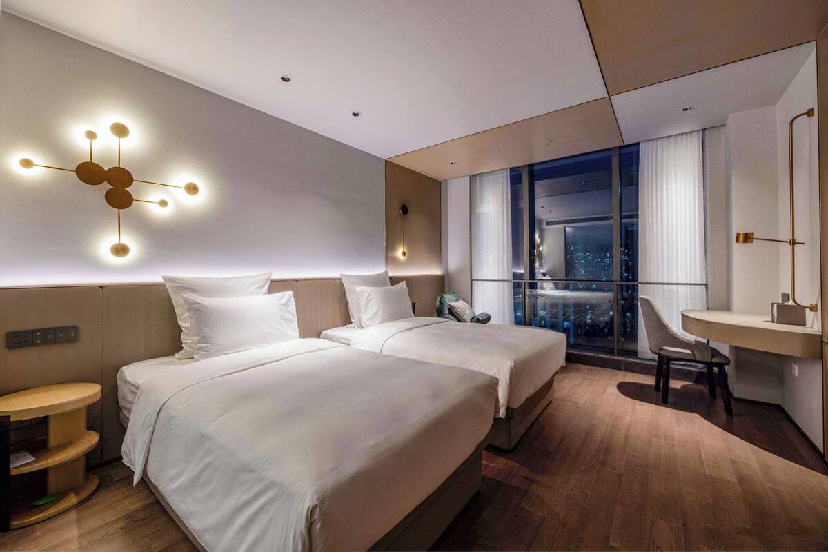 A modern hotel room with two beds, a desk, a chair, and a city view.