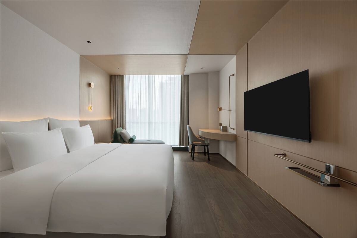 A modern hotel room with a large bed, a desk, a flat-screen TV, and a large window with curtains.