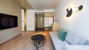 A modern living room with a flat-screen TV, a round coffee table, a grey sofa with a green pillow, and a wall-mounted light fixture. - Novotel Kunshan East (Suzhou)