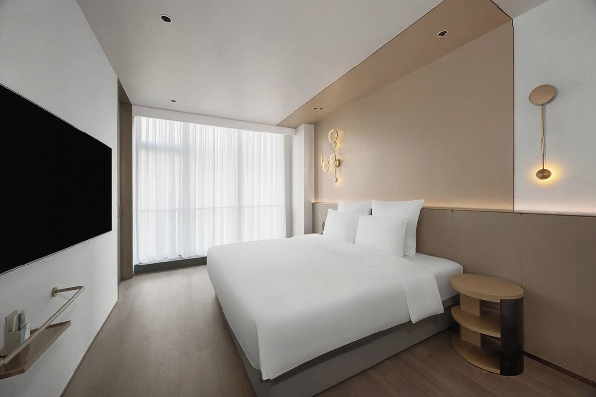 A modern hotel room with a large bed, a flat-screen TV, a bedside table, and a wall-mounted light fixture.