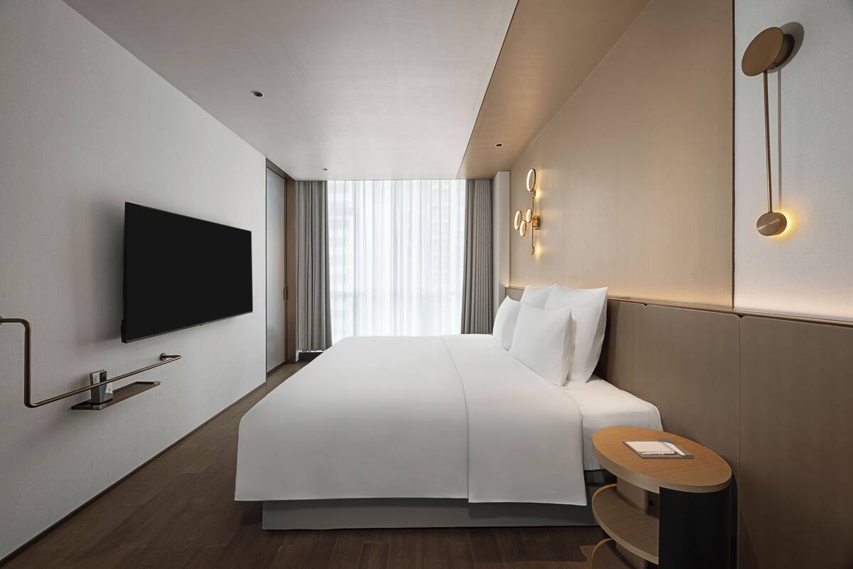 A modern hotel room with a large bed, a flat-screen TV, a bedside table, and a wall-mounted light fixture.