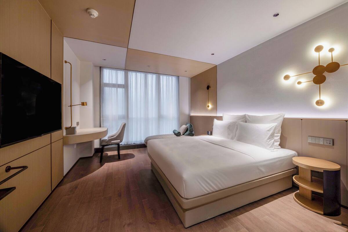A modern hotel room with a large bed, a TV, a desk, and a wall-mounted light fixture.