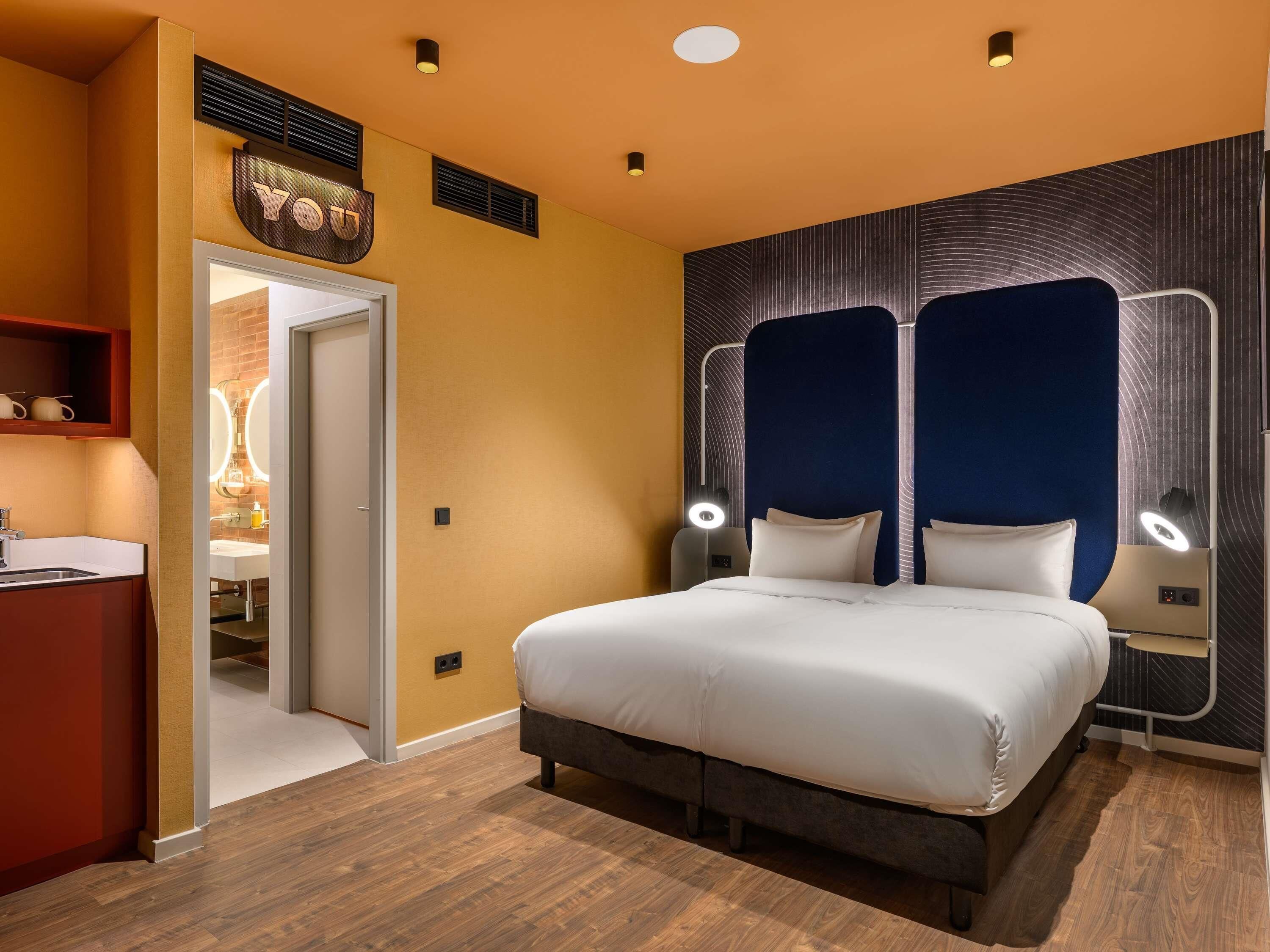 A hotel room with a bed, bedside lamps, a vanity with a mirror, and a sign above the door.
