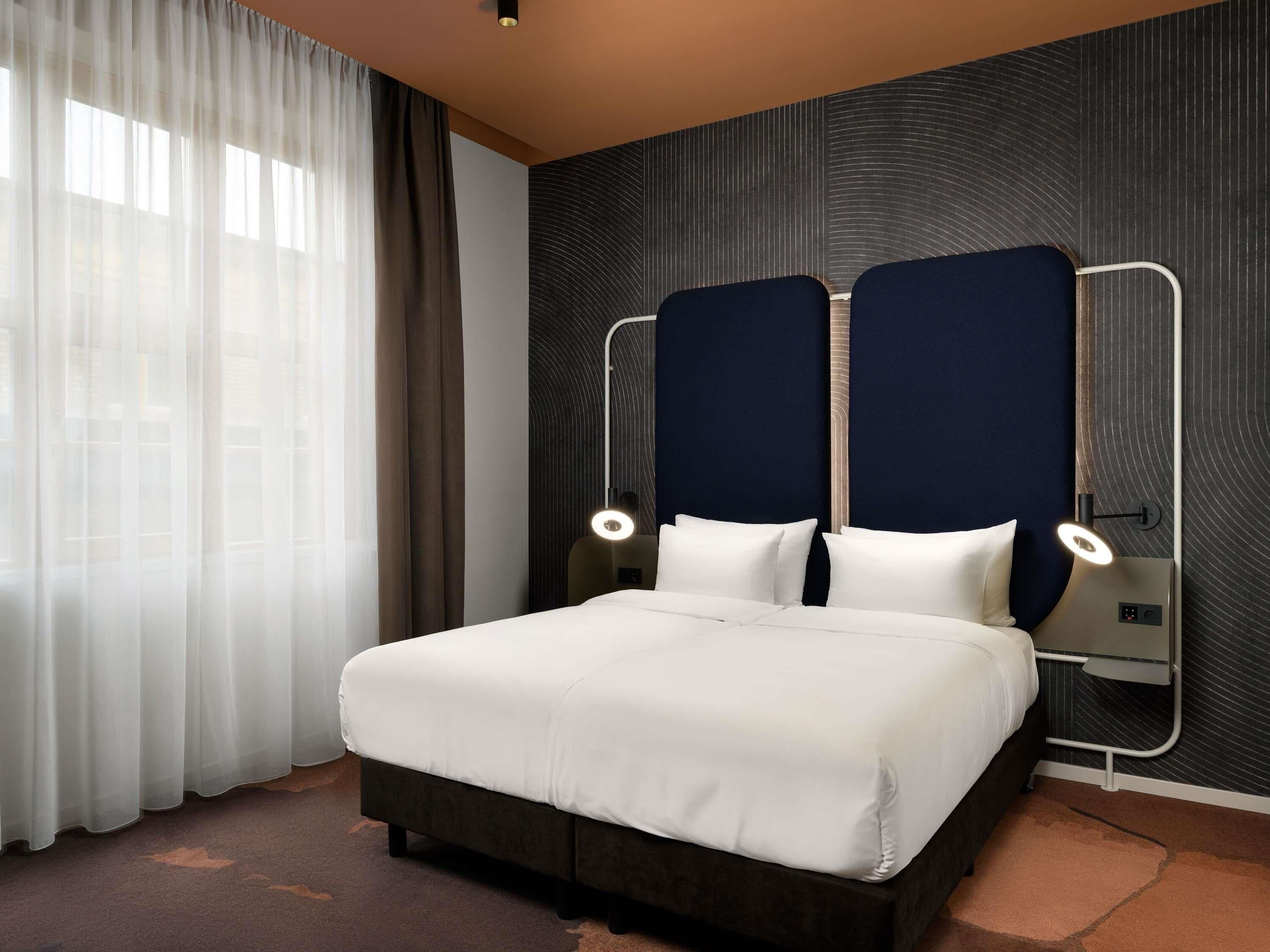 Habitación de hotel con una cama grande, dos mesitas de noche, un cabecero y una ventana con cortinas.