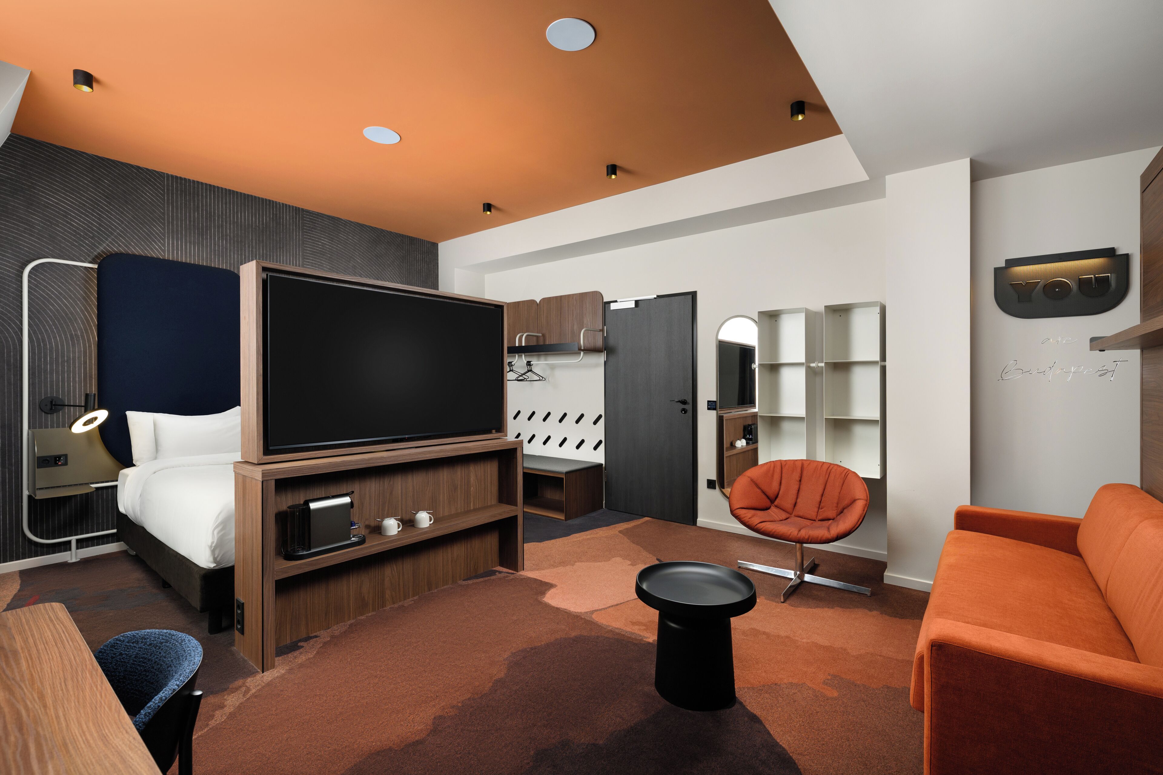 Una habitación de hotel moderna con una televisión de pantalla plana grande, una cama, un área de descanso con una silla y un sofá, una pequeña mesa y estanterías empotradas.