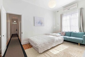 A bedroom with a bed, a sofa, a rug, a painting, and an air conditioning unit. - Oakley Cottage: CBD, Parking, Family Friendly (Adelaide)