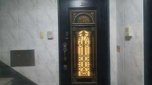 Une porte noire ornée d’un panneau décoratif doré, un extincteur et un escalier sur la gauche.