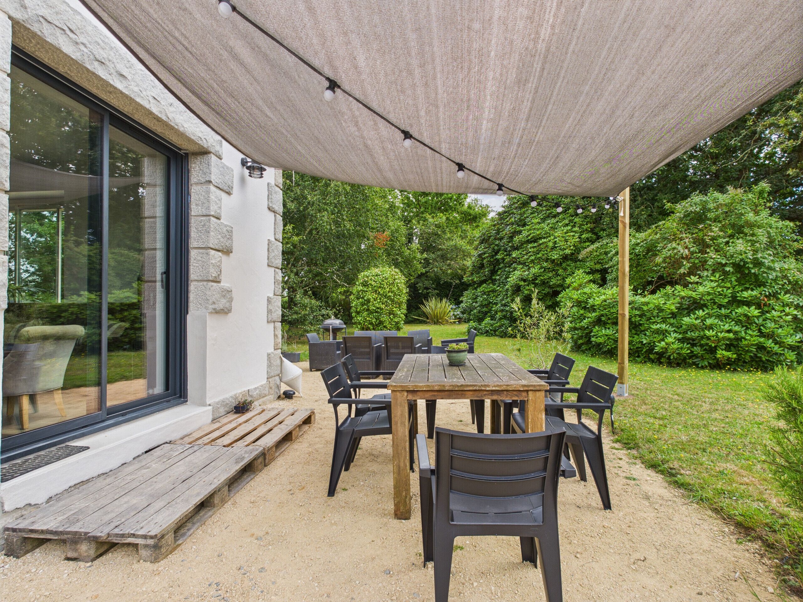 An outdoor patio with a table and chairs, a pergola providing shade, and a view of a garden with a hedge and trees.