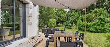 An outdoor patio with a table and chairs, a pergola providing shade, and a view of a garden with a hedge and trees.