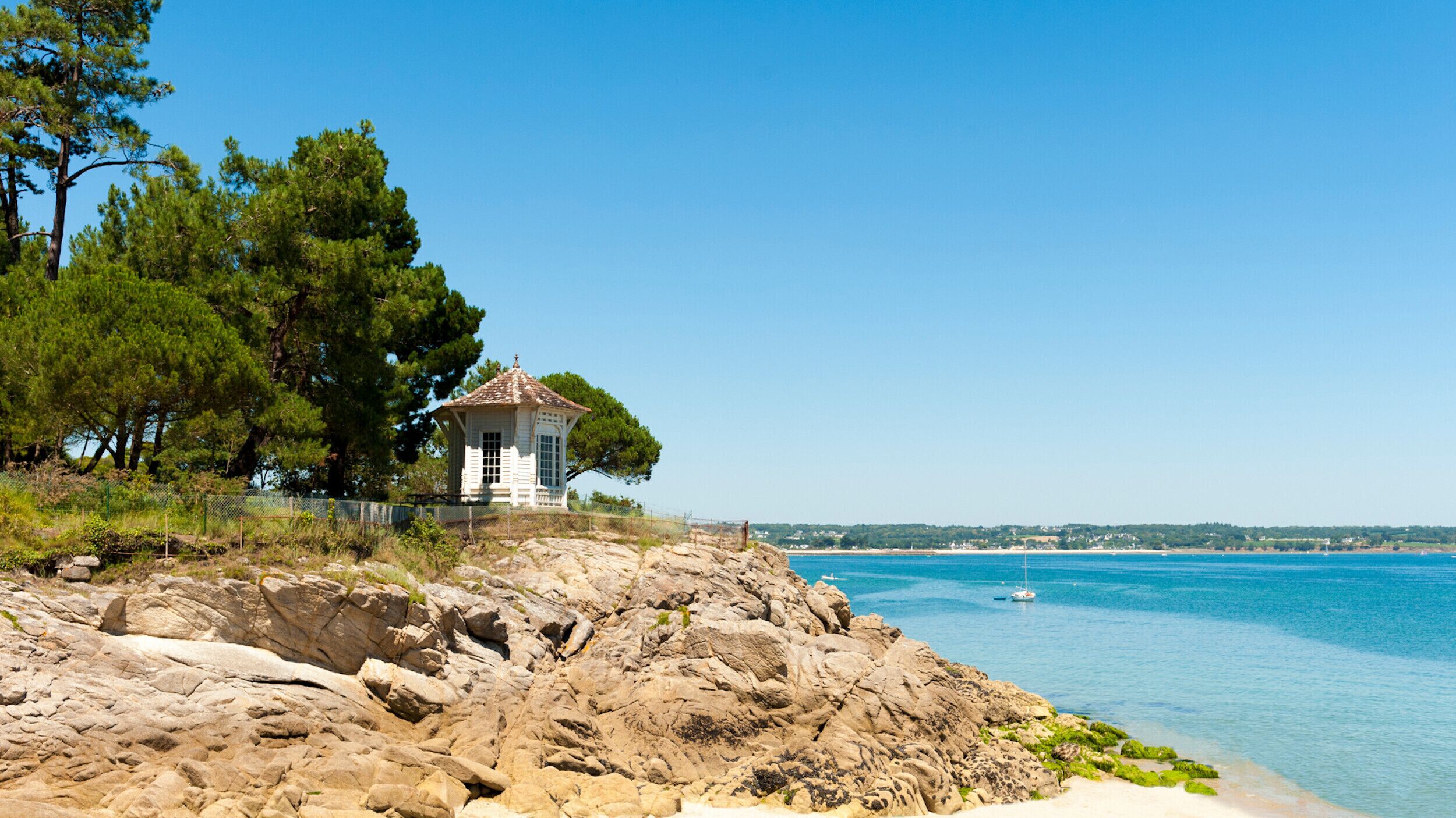 A coastal scene with a small white gazebo on rocky terrain, overlooking a calm sea and a distant shoreline.