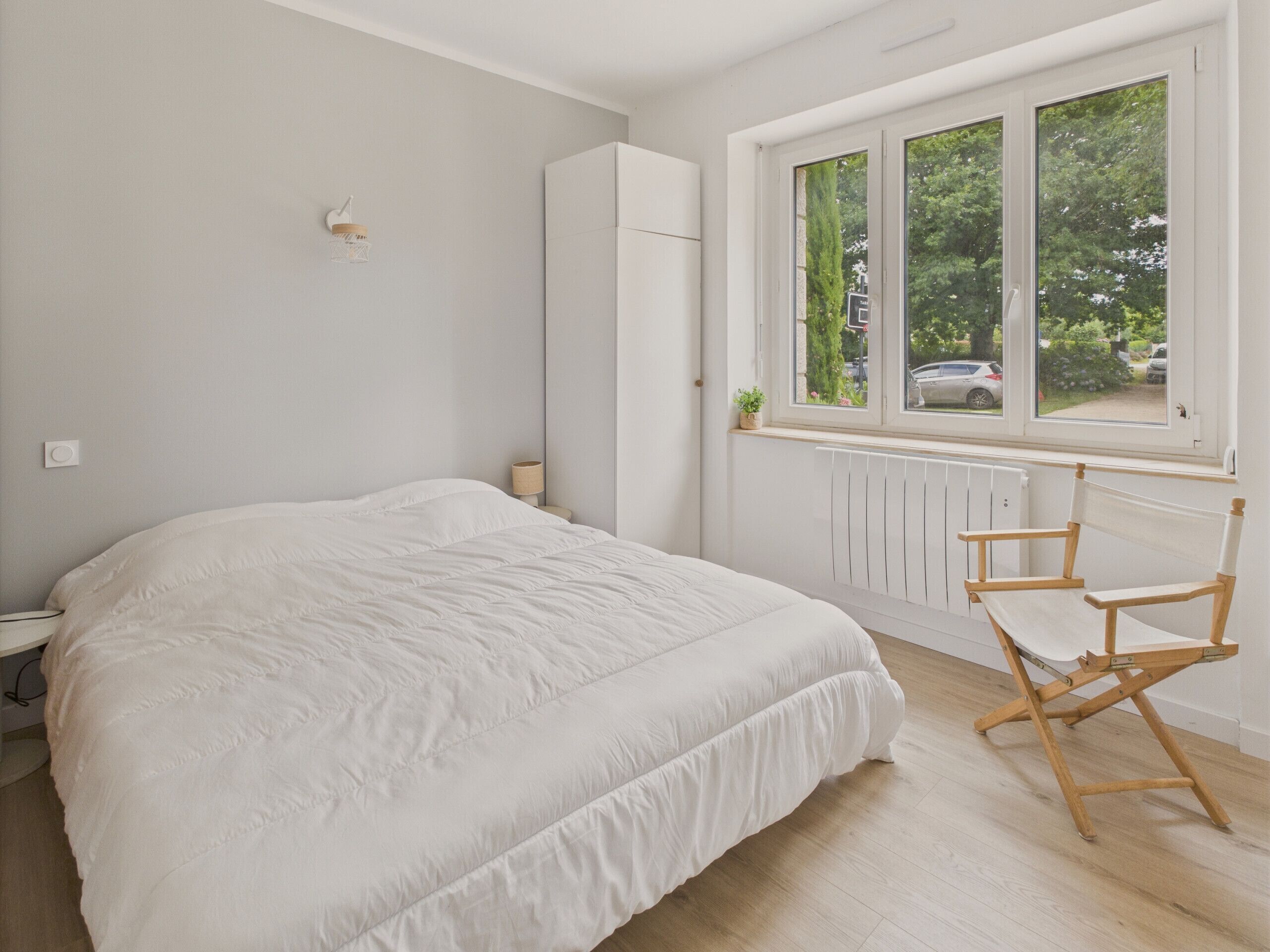 A bedroom with a large bed, a window with a view of the outside, a radiator, and a wooden chair.