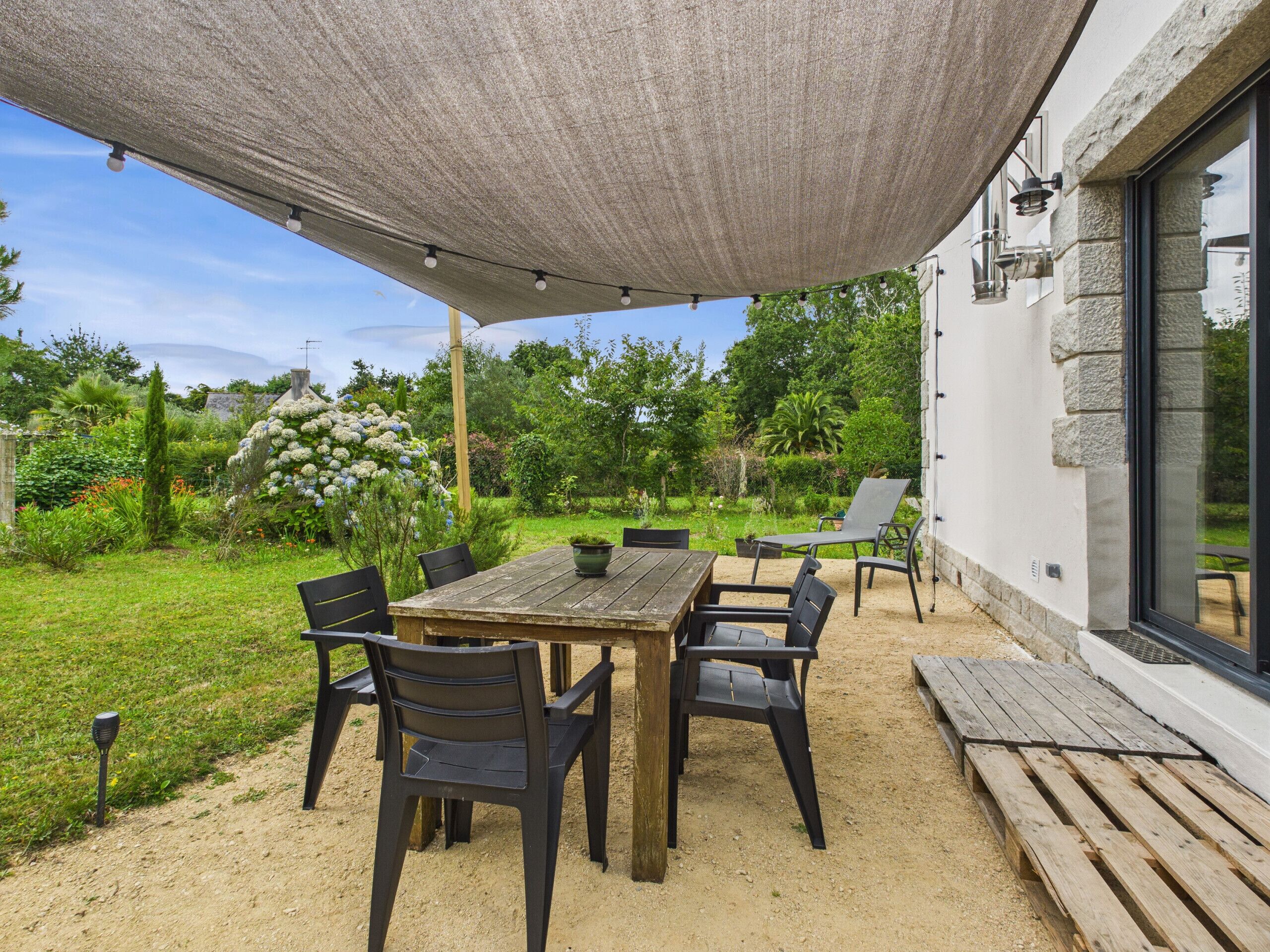 An outdoor patio with a wooden table and chairs, surrounded by a garden and a white building with stone accents.