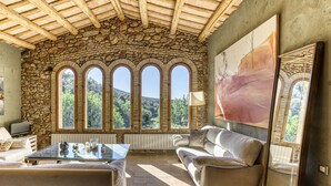 A room with stone walls, a stone ceiling, a large window with multiple panes, a sofa, a table, and a painting.