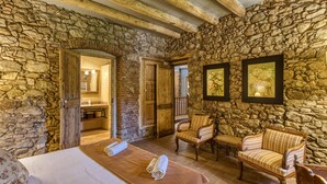 A stone-walled room with a bed, two chairs, and a bench. There are framed pictures on the wall and a wooden door leading to another room.