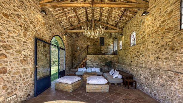 A stone-walled room with a wooden ceiling, a chandelier, and a seating area with cushions and a coffee table.