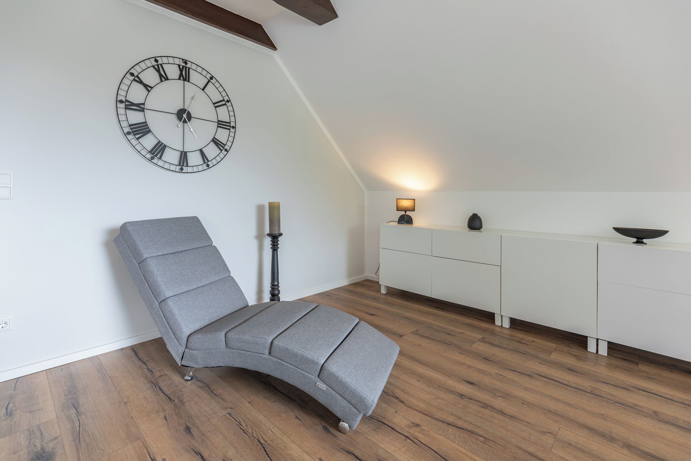 Une pièce moderne avec un canapé gris aux lignes courbes, une grande horloge murale, une petite table avec une lampe et un meuble blanc.