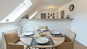 A dining area with a round table set for four, including plates, cutlery, and glasses. There are two chairs, a ceiling light, and a clock on the wall. - Vacation apartment "Heimathafen" in Breklum for up to 4 people (Breklum)