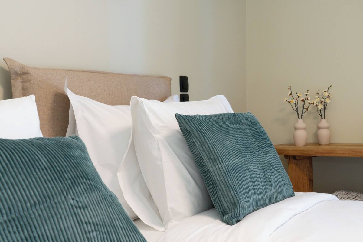 Una cama con almohadas blancas y turquesas, una mesita de noche de madera con dos jarrones de flores y una pared color beige.