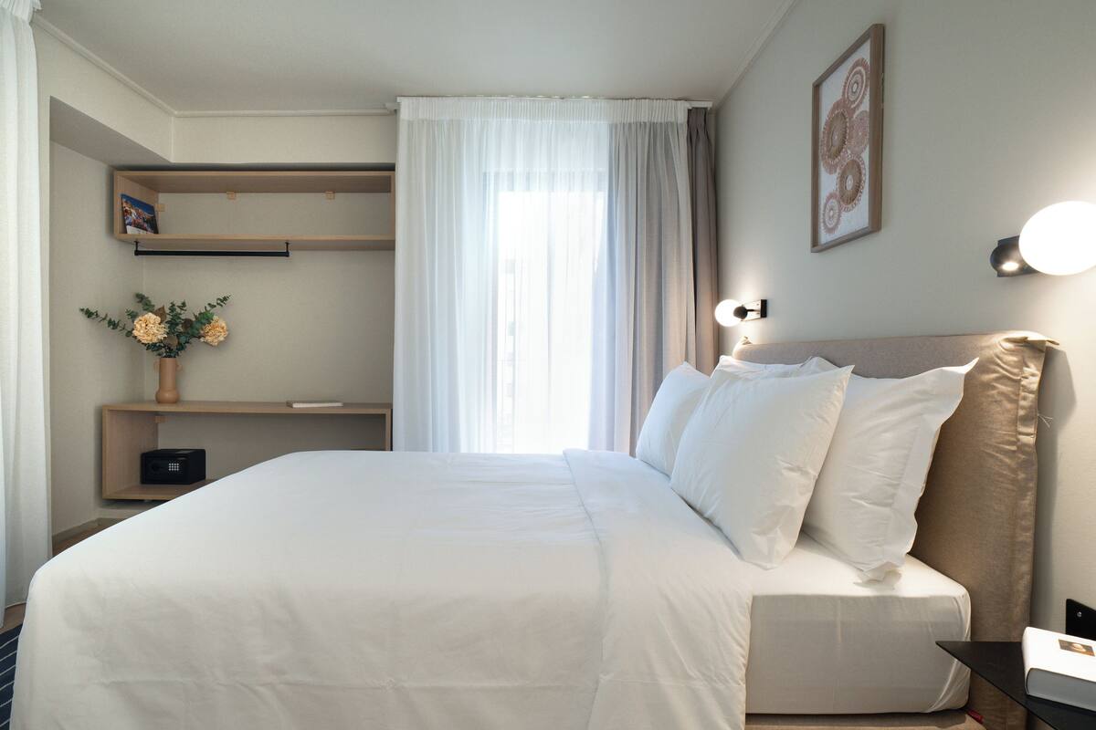 Una cama bien hecha con sábanas blancas, una mesita de noche con una lámpara, un estante con un jarrón de flores y una ventana con cortinas de encaje.