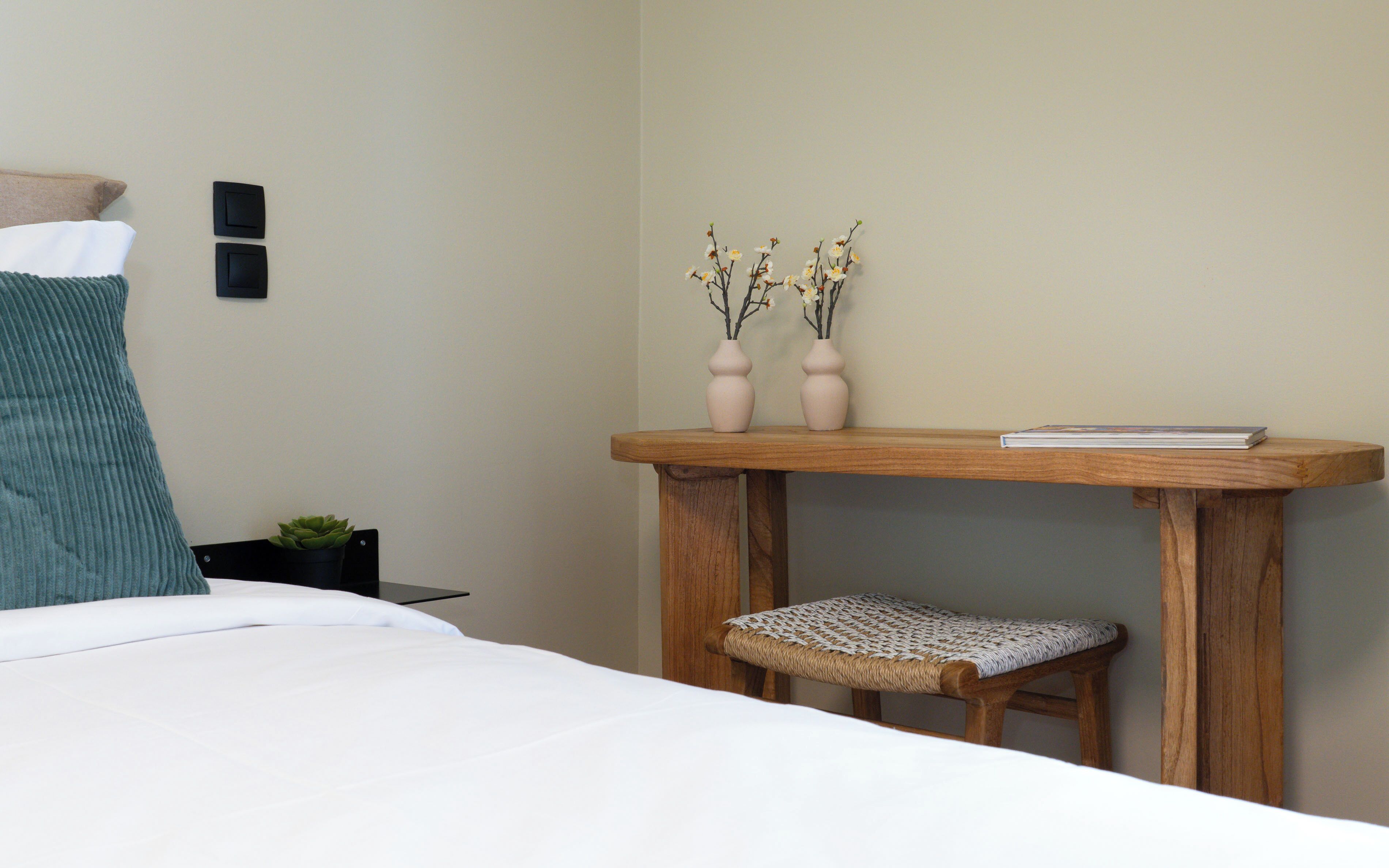 Ein Schlafzimmer mit einem Bett, einem kleinen Holztisch mit zwei Vasen und einem Buch, einem Korbstuhl und einem an der Wand befestigten Lichtschalter.
