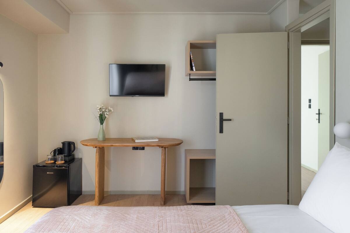 Habitación de hotel con cama, televisor montado en la pared, una mesita con un jarrón de flores, cafetera y puerta que da a otra habitación.