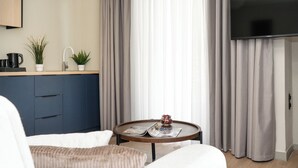 A modern hotel room with a flat-screen TV, a round wooden table with magazines, a sofa, a kitchen area with a sink and plants, and large curtains. - Gnamma Hotel (Palaio Faliro)