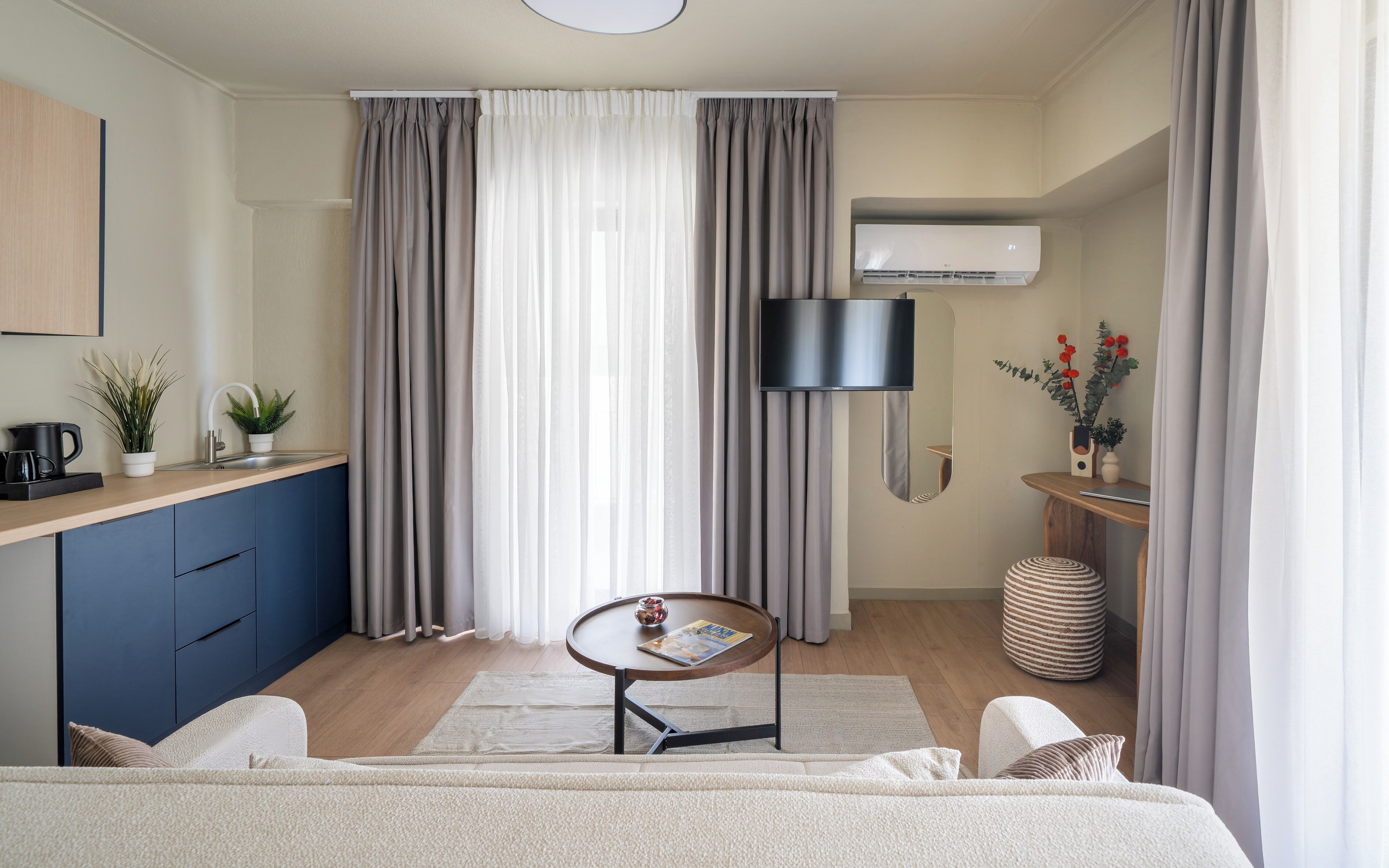 Una habitación de hotel moderna con una cama, una mesita redonda pequeña, un televisor montado en la pared, un lavamanos y un ventanal con cortinas.
