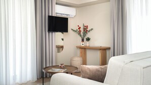 A modern living room with a flat-screen TV mounted on the wall, a round wooden coffee table, a beige sofa, and a small side table with decorative items. - Gnamma Hotel (Palaio Faliro)