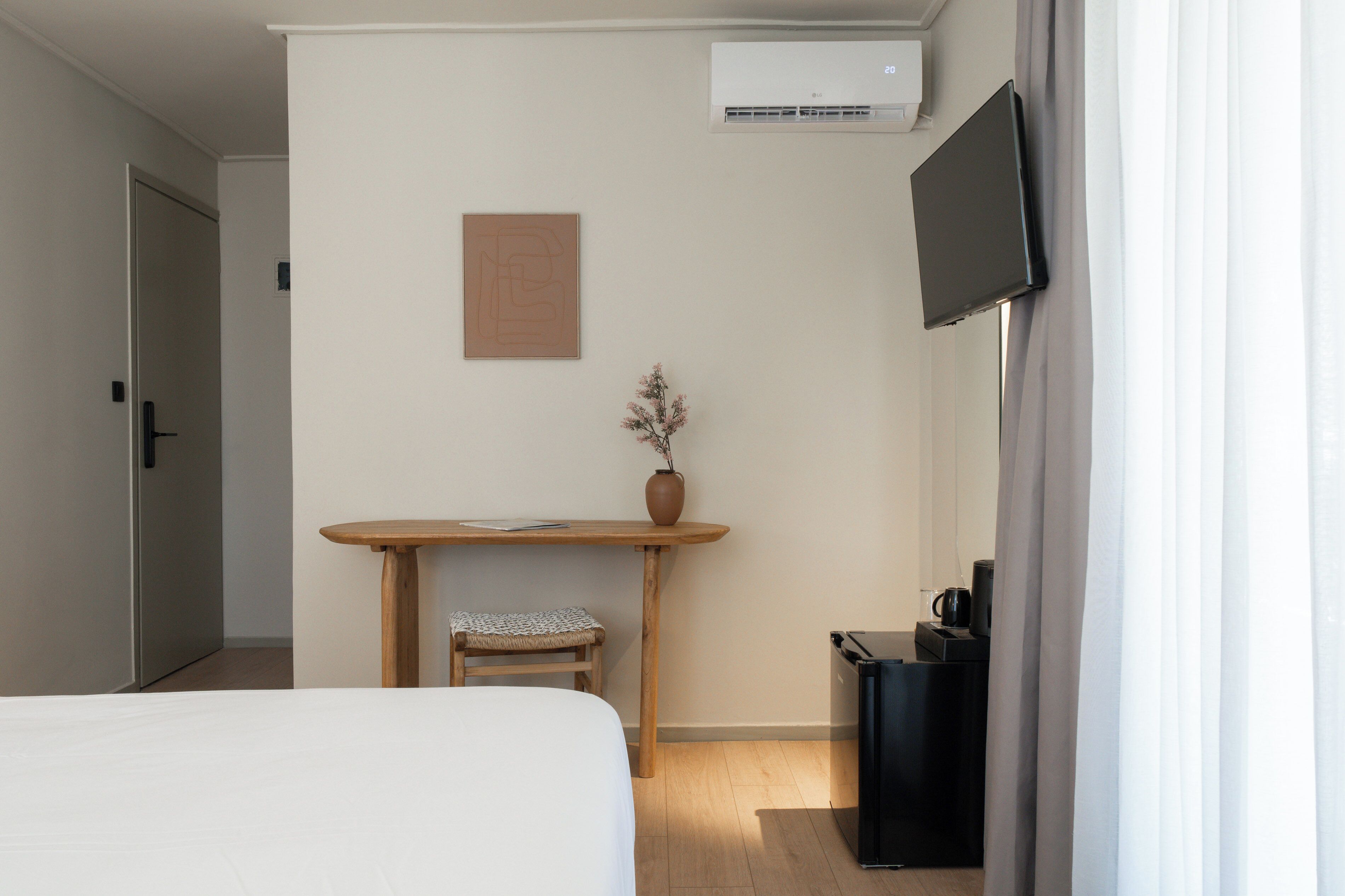 A hotel room with a bed, a desk with a chair, a small table, a vase with flowers, a television, and a trash bin.