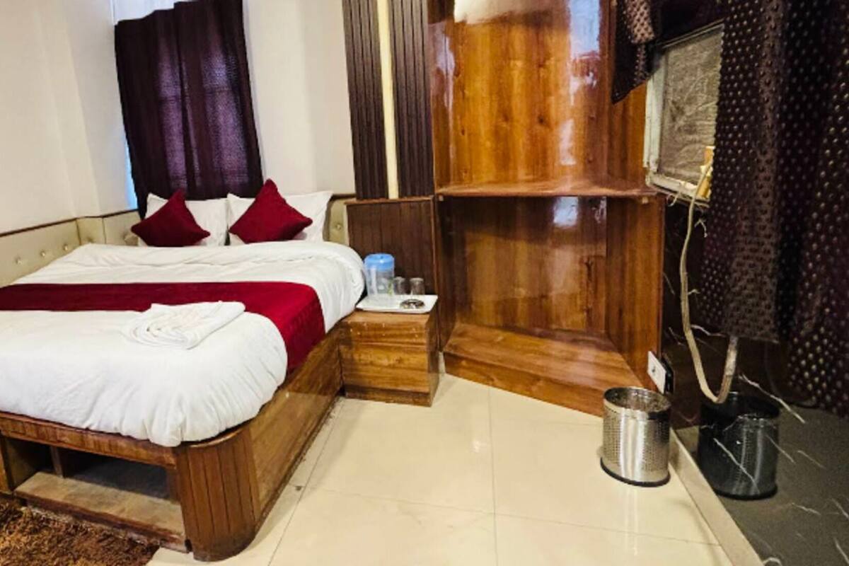 Cama bem arrumada com roupa de cama branca e vermelha, cabeceira de madeira, uma pequena mesa de cabeceira com bandeja e um cesto de lixo de metal.