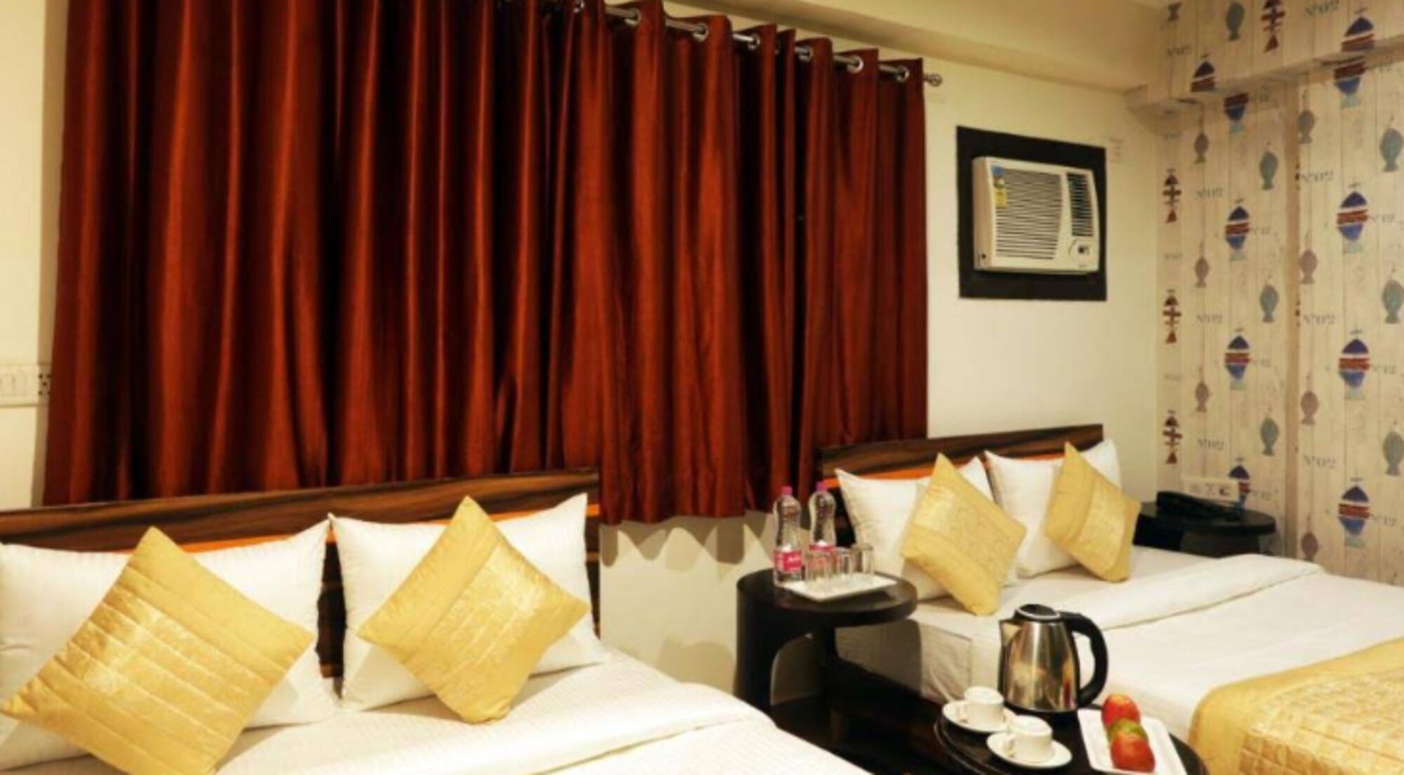 A hotel room with two beds, red curtains, a small table with a kettle, cups, and a tray with fruit.