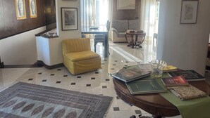Een hotelkamer met een bank, een gele fauteuil, een eettafel met boeken, een kamerbreed tapijt en een kroonluchter.