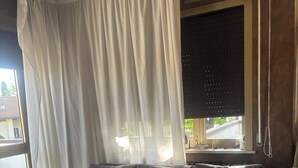 Een moderne woonkamer met een grijze bank, een groot raam met zonwering en een betegelde vloer.