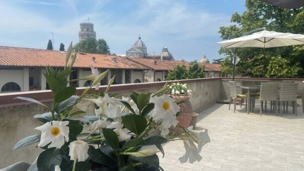Een terras op het dak met uitzicht op historische gebouwen, waaronder een toren en gebogen constructies. Er staan potplanten en is er buitenmeubilair.