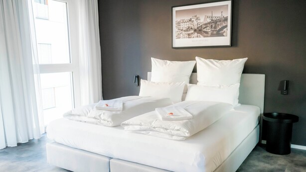 Een moderne hotelkamer met een groot bed, witte lakens, een ingelijst stadsgezicht aan de muur en een raam met gordijnen.