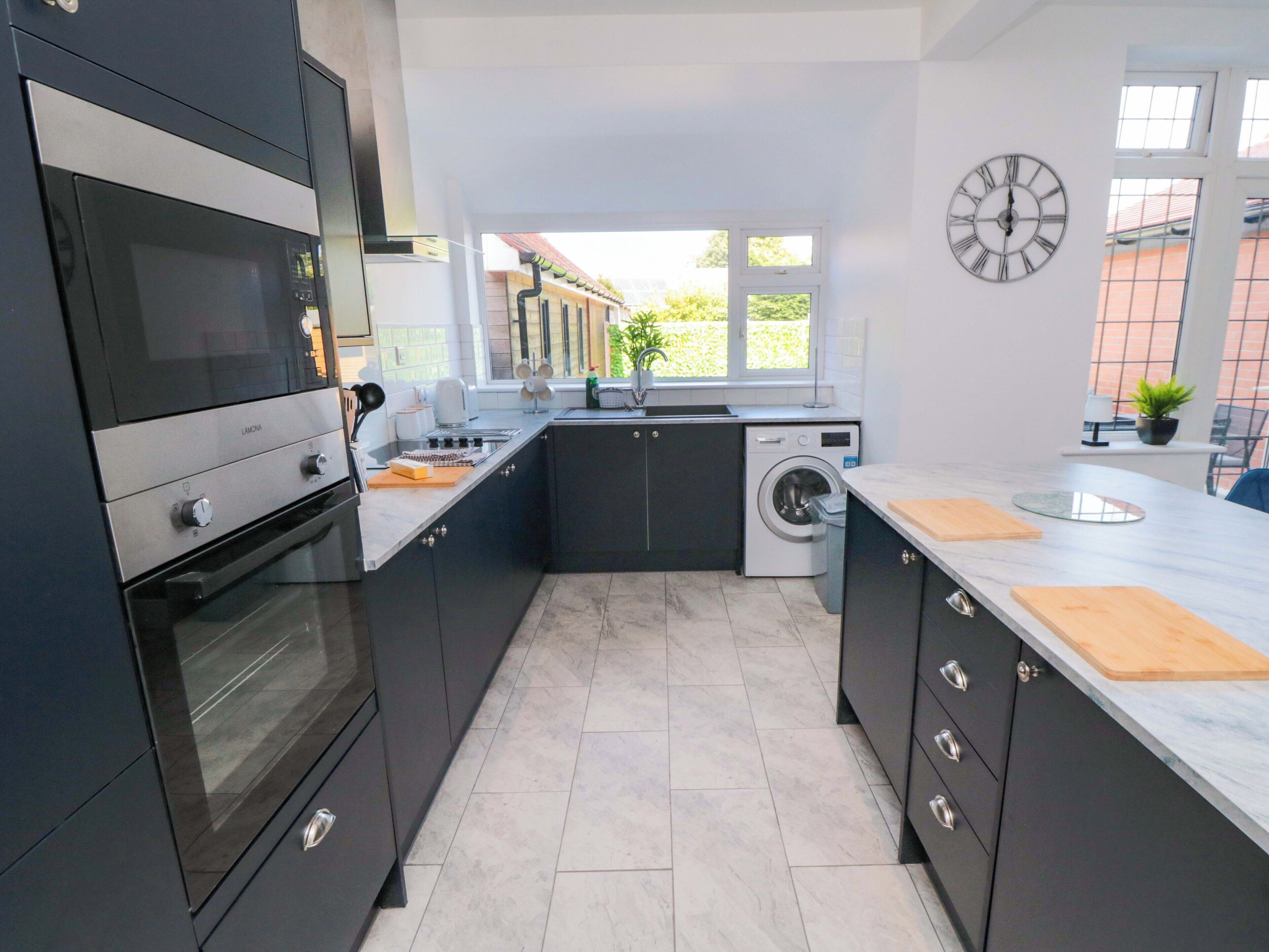 A modern kitchen with dark cabinets, a built-in oven, a washing machine, and a clock on the wall.