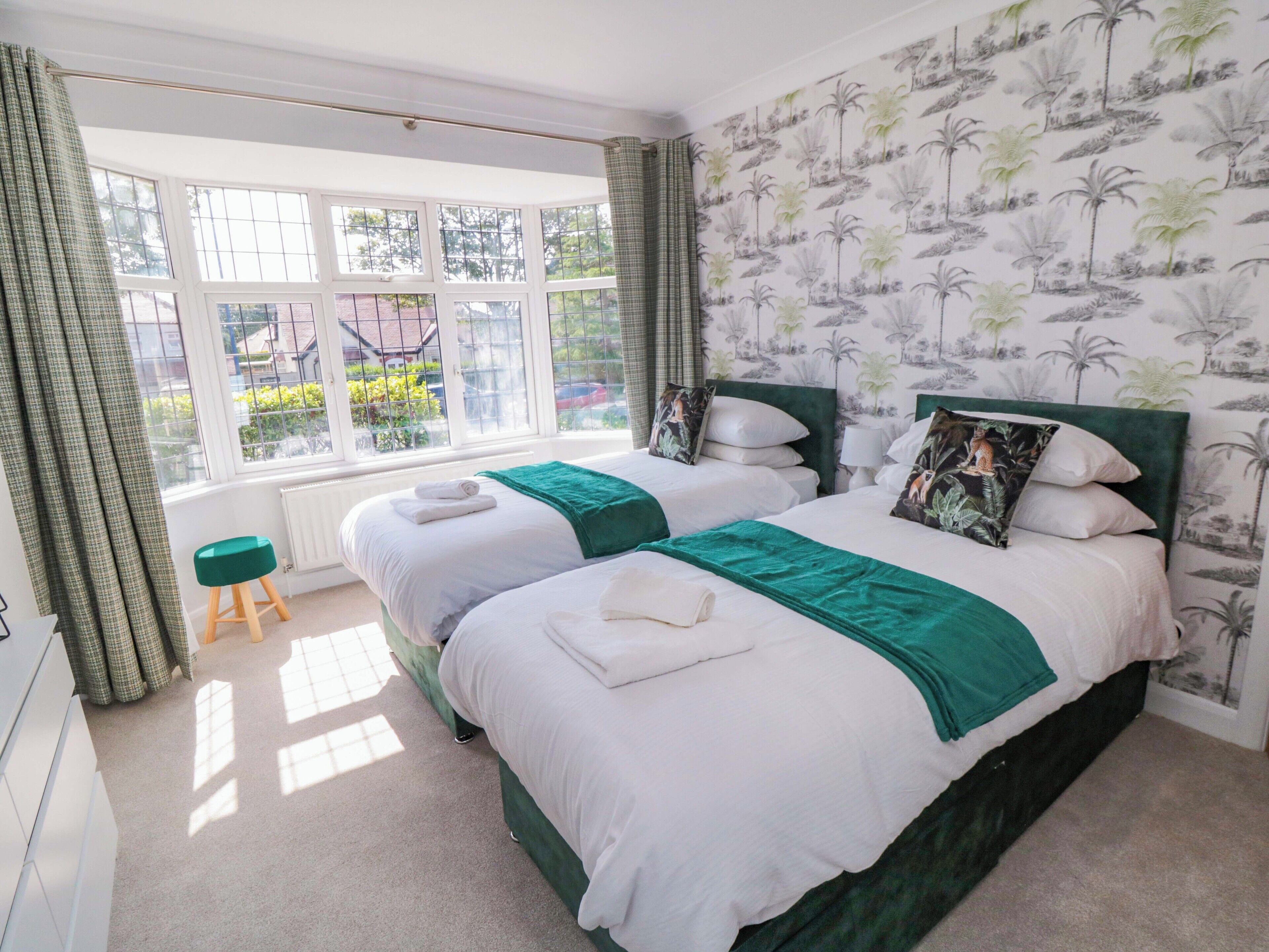 A double bed with a green headboard, a window with curtains, and a tropical wallpaper.